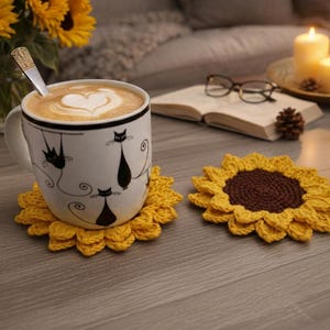 Op de afbeelding: Een witte mok met zwarte kattenillustraties staat op een gele gehaakte zonnebloemonderzetter. De mok bevat een latte met een hartvormig ontwerp. Er ligt nog een zonnebloemonderzetter naast de mok.