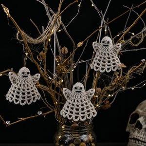 May include: Three beige crocheted ghost ornaments with black bead eyes and stitched mouths, hanging from thin strings on bare branches. Each ghost features a wide, flared skirt with an openwork lattice pattern and small, outstretched arms.