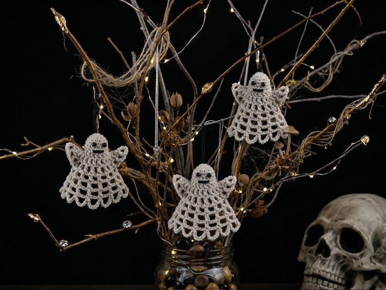 May include: Three beige crocheted ghost ornaments with black bead eyes and stitched mouths, hanging from thin strings on bare branches. Each ghost features a wide, flared skirt with an openwork lattice pattern and small, outstretched arms.
