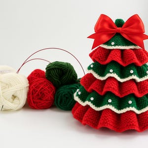 K&ouml;nnte beinhalten: Handgefertigte Weihnachtsbaumdekoration mit roter Schleife. Der Baum besteht aus roter und gr&uuml;ner Strickwolle, mit wei&szlig;em Rand und Perlenakzenten. Daneben liegen Wollkn&auml;uel in Wei&szlig;, Rot und Gr&uuml;n, was auf ein Bastelprojekt hindeutet.
