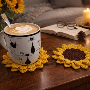 May include: A white mug with black cat illustrations, filled with coffee and a heart-shaped design in the foam, sits on a yellow crochet sunflower coaster. Another sunflower coaster is next to it. A vase of sunflowers and candles are in the background.