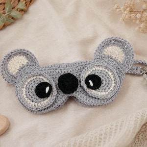 May include: A handmade gray crochet sleep mask shaped like a koala. The mask features black eyes, white accents, and a strap with a metal clasp. The ears are also gray with white centers. The mask is designed for sleep and relaxation.