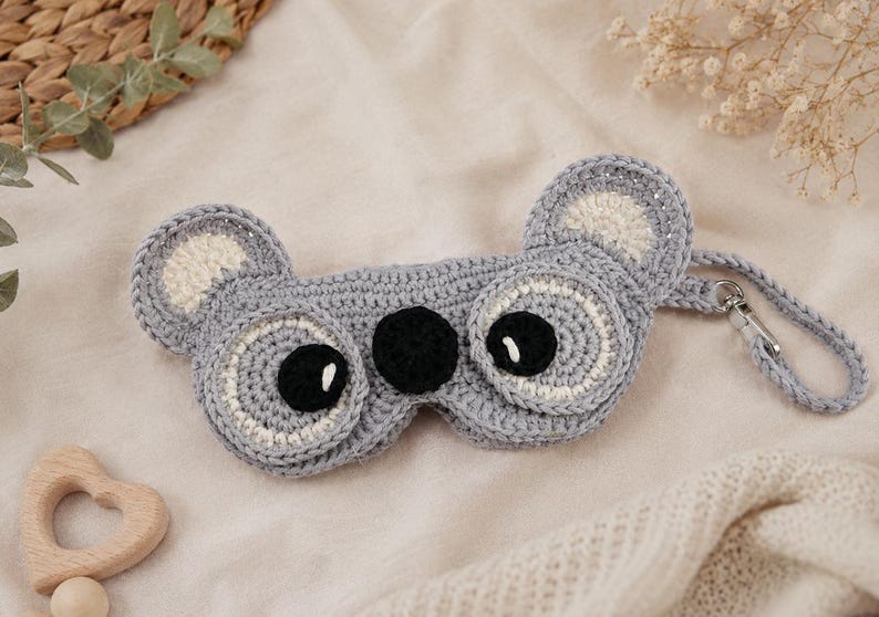 May include: A handmade gray crochet sleep mask shaped like a koala. The mask features black eyes, white accents, and a strap with a metal clasp. The ears are also gray with white centers. The mask is designed for sleep and relaxation.