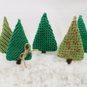 May include: Five knitted Christmas tree decorations in various shades of green. Some trees have red beads for decorations. The trees are sitting on a white fluffy surface.