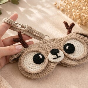 May include: A crocheted sleep mask in the shape of a deer, featuring large, round eyes with black pupils and a small black nose. The mask is beige with brown antlers and a matching strap with a metal clasp.