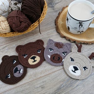 May include: Four handmade crochet coasters in the shape of animal faces: two bears, a boar, and a deer. The coasters are brown, gray, and tan with black and white details. A basket of yarn and a mug of coffee sit nearby.