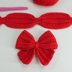 Puede incluir: Primer plano de lazos y una diadema hechos a mano de ganchillo rojo. Tambi&eacute;n se ven un ganchillo rosa y una madeja de hilo rojo. Los lazos y la diadema est&aacute;n sobre una superficie blanca.
