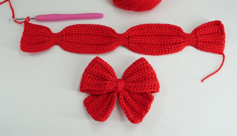 Puede incluir: Primer plano de lazos y una diadema hechos a mano de ganchillo rojo. Tambi&eacute;n se ven un ganchillo rosa y una madeja de hilo rojo. Los lazos y la diadema est&aacute;n sobre una superficie blanca.