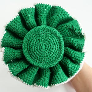 May include: A handmade, green crochet item with a white trim. The circular design features a central disc surrounded by ruffled, petal-like layers. The item is held against a white background.