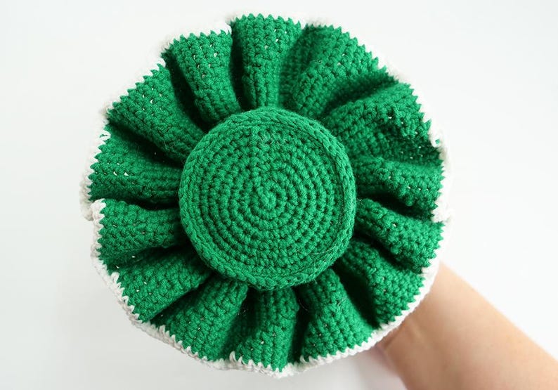 May include: A handmade, green crochet item with a white trim. The circular design features a central disc surrounded by ruffled, petal-like layers. The item is held against a white background.