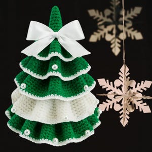 May include: A handmade, green and white crocheted Christmas tree with a white bow at the top. The tree is decorated with faux pearls and sits in front of two hanging snowflake ornaments. The background is dark.