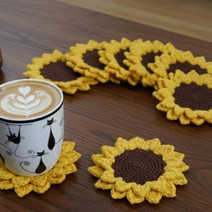 May include: A white mug with black cat illustrations sits on a yellow crochet sunflower coaster. Several more sunflower coasters are arranged on a wooden surface. The coasters have a brown center and yellow petals. A cup of coffee with latte art is in the mug.