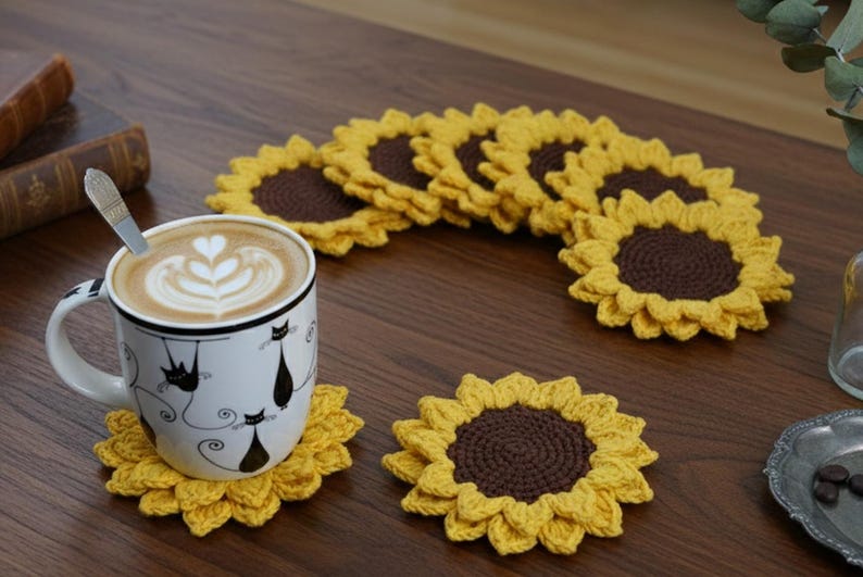 May include: A white mug with black cat illustrations sits on a yellow crochet sunflower coaster. Several more sunflower coasters are arranged on a wooden surface. The coasters have a brown center and yellow petals. A cup of coffee with latte art is in the mug.