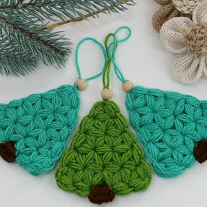 Pode incluir: Três enfeites de árvore de Natal feitos à mão em crochê. Dois são verde-azulado e um é verde. Cada árvore tem um tronco marrom e uma conta de madeira no topo. Os enfeites são exibidos em uma superfície branca com um galho de sempre-viva e flores de tecido.