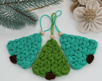 Puede incluir: Tres adornos de árbol de Navidad hechos a mano con ganchillo. Dos son de color verde azulado y uno es verde. Cada árbol tiene un tronco marrón y una cuenta de madera en la parte superior. Los adornos se exhiben sobre una superficie blanca con una ramita de hoja perenne y flores de tela.