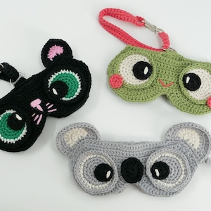 May include: Crocheted eye masks in the shape of a black cat, a green frog, and a gray koala. Each mask has a strap and is detailed with colorful accents. The cat has pink ears and whiskers, the frog has pink cheeks, and the koala has white ears.