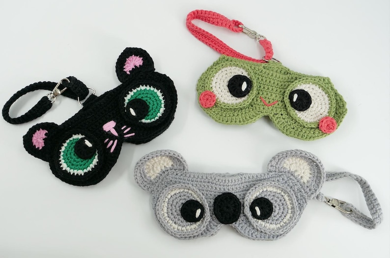 May include: Crocheted eye masks in the shape of a black cat, a green frog, and a gray koala. Each mask has a strap and is detailed with colorful accents. The cat has pink ears and whiskers, the frog has pink cheeks, and the koala has white ears.