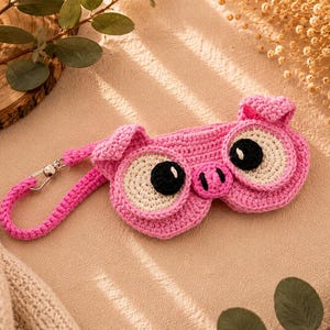 May include: A pink crocheted sleep mask shaped like a pig face, featuring large round eyes with black pupils, a small snout, and two pointed ears. The mask includes a matching pink braided strap with a silver-toned metal lobster clasp attachment.