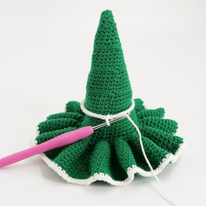 May include: A green crocheted Christmas tree with a white trim and a pink crochet hook. The tree is in the process of being crocheted. The tree has a cone shape and a ruffled base.