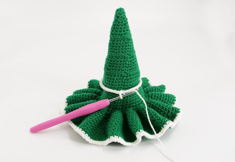May include: A green crocheted Christmas tree with a white trim and a pink crochet hook. The tree is in the process of being crocheted. The tree has a cone shape and a ruffled base.