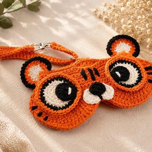 May include: An orange crochet eyeglass case shaped like a tiger's face, featuring black and white details for the eyes, nose, and ears. The case has a matching strap with a silver clasp. The case is handmade.