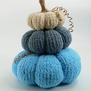 Op de afbeelding: Een stapel van drie pluche pompoenen in tinten blauw en beige. De onderste pompoen is lichtblauw, de middelste is donkerder blauw en de bovenste is beige. Elke pompoen heeft een getextureerd, geribbeld oppervlak en een bruine stengel.