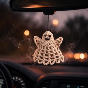 May include: A cream-coloured crocheted ghost ornament hanging from a black string. The ghost features a rounded head, two small black bead eyes, a straight black stitched mouth, and a wide, flared body with a scalloped bottom edge design.
