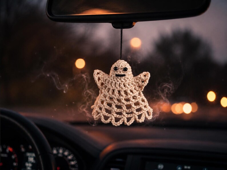 May include: A cream-coloured crocheted ghost ornament hanging from a black string. The ghost features a rounded head, two small black bead eyes, a straight black stitched mouth, and a wide, flared body with a scalloped bottom edge design.