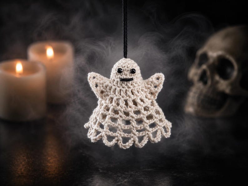 May include: A small, off-white crocheted ghost ornament featuring a bell-shaped body with an open-work lattice pattern, two small arms extended outwards, and two black bead eyes with a stitched black mouth, suspended by a thin black string.