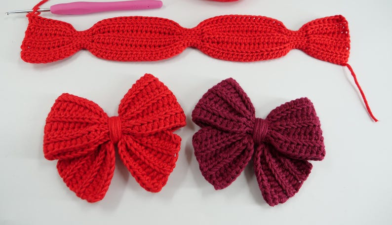Puede incluir: Lazos de ganchillo hechos a mano en rojo y burdeos, con una textura acanalada. La imagen muestra lazos en diferentes etapas de finalizaci&oacute;n, junto con un ganchillo rosa e hilo rojo. Artesan&iacute;a.