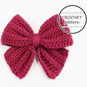 Puede incluir: Lazo de ganchillo hecho a mano, de color magenta. El lazo est&aacute; hecho con un hilo grueso y presenta un patr&oacute;n de puntadas texturizadas. El texto "CROCHET pattern" est&aacute; en un c&iacute;rculo negro en la esquina superior derecha. El lazo est&aacute; centrado sobre un fondo blanco.