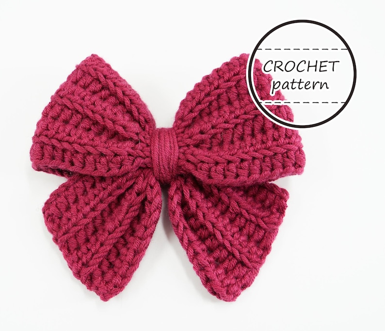 Puede incluir: Lazo de ganchillo hecho a mano, de color magenta. El lazo est&aacute; hecho con un hilo grueso y presenta un patr&oacute;n de puntadas texturizadas. El texto "CROCHET pattern" est&aacute; en un c&iacute;rculo negro en la esquina superior derecha. El lazo est&aacute; centrado sobre un fondo blanco.