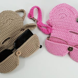 May include: Two crochet eyeglass cases in the image. One is tan, open, and holding black sunglasses. The other is pink, closed, and holding eyeglasses. A pink lanyard is attached to the pink case. Both cases have a button closure.