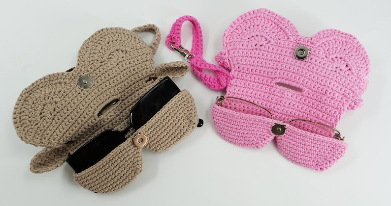 May include: Two crochet eyeglass cases in the image. One is tan, open, and holding black sunglasses. The other is pink, closed, and holding eyeglasses. A pink lanyard is attached to the pink case. Both cases have a button closure.