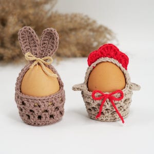Op de afbeelding: Twee gehaakte eierhouders. Eén is bruin met konijnenoren en een touwstrik. De andere is beige met een rode kam en een rode strik. Beide houders omvatten bruine eieren, perfect voor Pasen.
