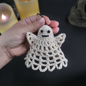 May include: A small, off-white crocheted ghost ornament featuring a delicate open-weave pattern, two small black bead eyes, and a simple stitched black mouth. The ghost has a rounded head, outstretched arms, and a scalloped bottom edge.