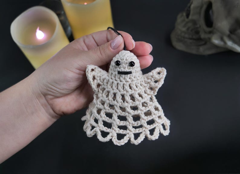 May include: A small, off-white crocheted ghost ornament featuring a delicate open-weave pattern, two small black bead eyes, and a simple stitched black mouth. The ghost has a rounded head, outstretched arms, and a scalloped bottom edge.