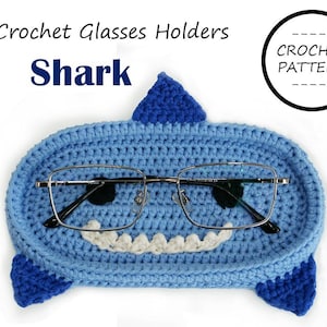 Puede incluir: Un soporte para gafas de ganchillo azul claro con forma de tiburón, con boca y dientes blancos. El soporte tiene una aleta en la parte superior y dos aletas laterales. El texto dice "Crochet Glasses Holders" y "Shark". Las gafas tienen montura plateada.