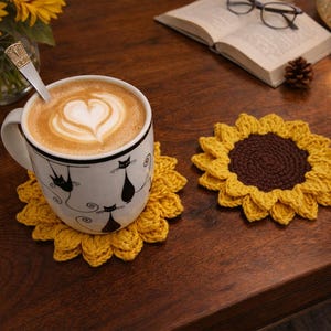 May include: A white mug with black cat silhouettes, filled with coffee and latte art, sits on a yellow crochet sunflower coaster. Another sunflower coaster is next to it. The scene is on a dark wooden table.