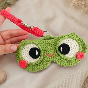 May include: A green crochet eye mask featuring large white and black eyes, a small red stitched mouth, and two pink circular cheeks. The mask is attached to a bright pink crochet wrist strap with a silver metal lobster clasp and ring.