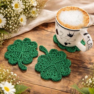 Può includere: Due sottobicchieri a forma di trifoglio all'uncinetto verde fatti a mano su una superficie di legno accanto a una tazza di caffè. La tazza ha un disegno di gatto nero ed è piena di una bevanda schiumosa. Un mazzo di fiori bianchi è sullo sfondo.