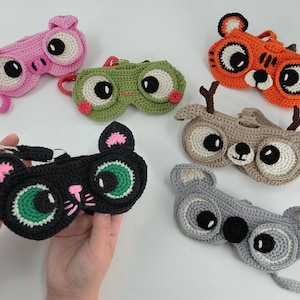 May include: A collection of handmade crochet eyeglass cases in various animal designs. The cases include a pink pig, green frog, orange tiger, brown deer, black cat, and gray koala. Each case features large, expressive eyes and a strap.