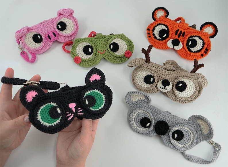May include: A collection of handmade crochet eyeglass cases in various animal designs. The cases include a pink pig, green frog, orange tiger, brown deer, black cat, and gray koala. Each case features large, expressive eyes and a strap.