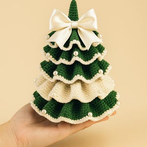 May include: A handcrafted, crochet Christmas tree in green and cream, adorned with faux pearls and a cream satin bow. The tree is held in a hand, showcasing its detailed, layered design. A festive, decorative item.