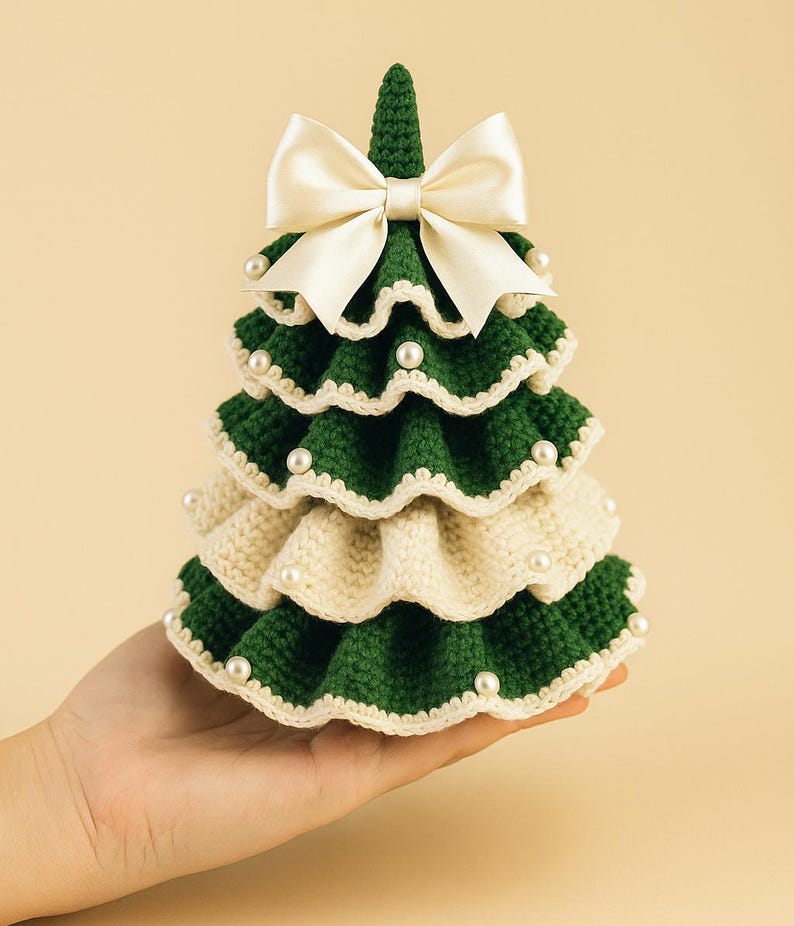 May include: A handcrafted, crochet Christmas tree in green and cream, adorned with faux pearls and a cream satin bow. The tree is held in a hand, showcasing its detailed, layered design. A festive, decorative item.