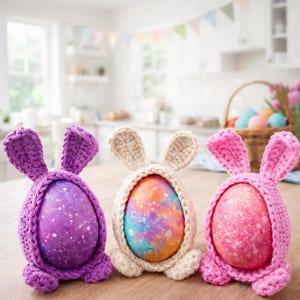 May include: Three decorative Easter eggs in handmade crochet bunny egg holders. The eggs are painted with speckled designs in purple, orange, and pink. The bunny holders are in matching colours, with upright ears.