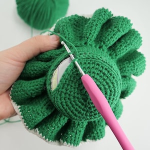 May include: A close-up of a green crocheted item with a white border, possibly a flower or a similar design. A pink crochet hook is inserted into the item, with a ball of green yarn in the background. The item is being crafted.