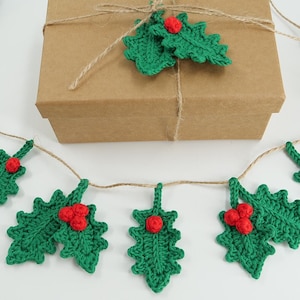 May include: A handmade Christmas garland featuring green crocheted holly leaves with red berry clusters. The garland is strung on twine and is displayed with a brown gift box tied with twine. A ball of green yarn is in the background.