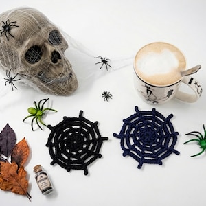 May include: A beige human skull covered in white spider web, two round crocheted spider web coasters in black and dark blue, a white mug with a cat graphic, a small glass bottle labeled poison, green plastic spiders, and dried autumn leaves.