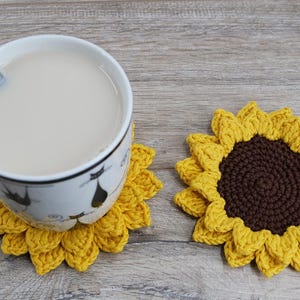 May include: A cup of tea with a spoon rests on a yellow crochet sunflower coaster. A second sunflower coaster is next to the cup. The coasters have a brown center and yellow petals. The cup has a black cat design.